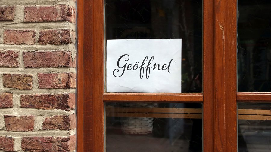 In einem Fenster mit Holzrahmen hängt ein Schild mit dem Hinweis „Geöffnet“.