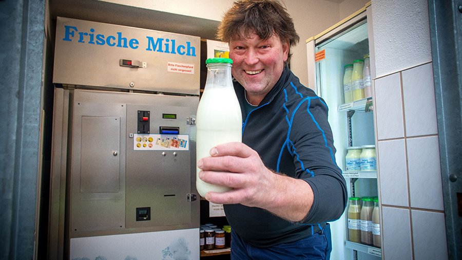 Ein Landwirt steht vor einer Milchtankstelle und hält eine Glasflasche mit abgezapfter Milch in die Kamera. 