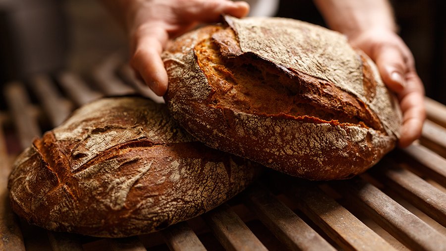 Zwei Hände legen einen Brotlaib zu einem anderen auf einen Holzrost.