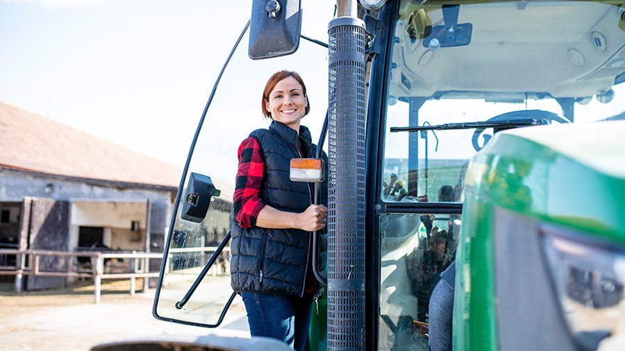 Frau steht auf Treppe eines grünen, neuen Traktors, schaut in Kamera. Bild: Halfpoint – stock.adobe.com