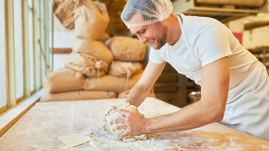 Ein junger Bäckergeselle steht an einem Tisch mit mehlbedeckter Arbeitsplatte und knetet Brotteig. Im Hintergrund liegen Mehlsäcke.