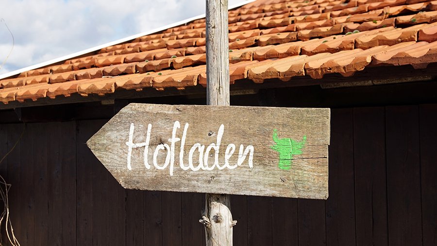 Ein Holzschild mit der Aufschrift „Hofladen“ ist an einem Holzpfosten angebracht und zeigt nach links. Es steht vor einem Schuppen rotem Ziegeldach. 