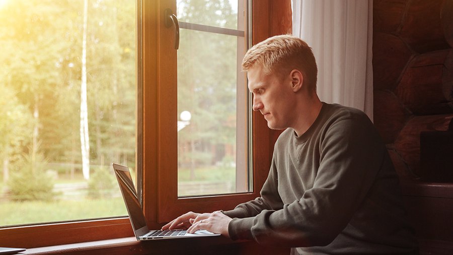 Ein junger Mann sitzt mit einem Laptop am Fenster mit Blick auf eine gründe und ländliche Umgebung.