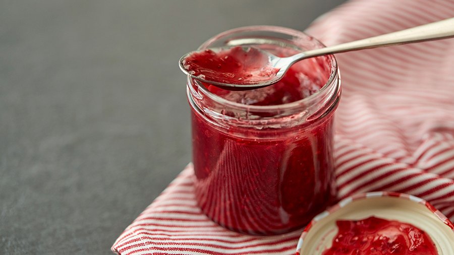 Ein geöffnetes Marmeladenglas steht auf einem rot-weiß gestreiften Küchenhandtuch, der Deckel nach oben geöffnet daneben. Auf dem Glas liegt ein Teelöffel.