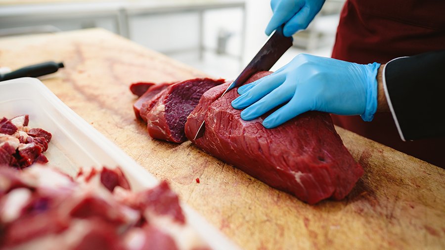 Eine Person mit blauen Gummi-Handschuhen zerschneidet ein Stück Fleisch auf einem Holzbrett.