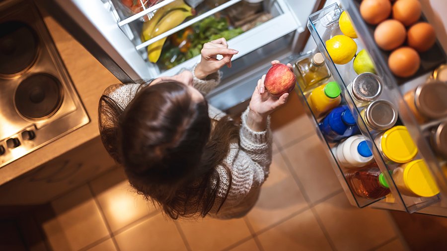 Von oben betrachtet man eine Frau, die vor einem geöffneten und gut gefüllten Kühlschrank steht und einen Apfel in der Hand hält.